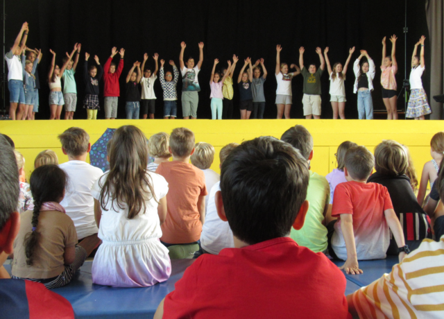 Im Hintergrund steht Klasse 4a auf einer gelben Bühne und streckt während eines Bewegungsliedes die Arme nach oben. Im Vordergrund sitzen Kinder, die zuschauen.