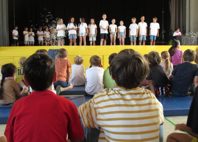 Auf einer gelben Bühne stehen die Kinder der 4b in Jeans und weißem T-Shirt und singen. Im Vordergrund des Bildes sitzen mehrere Kinder, die zuschauen.