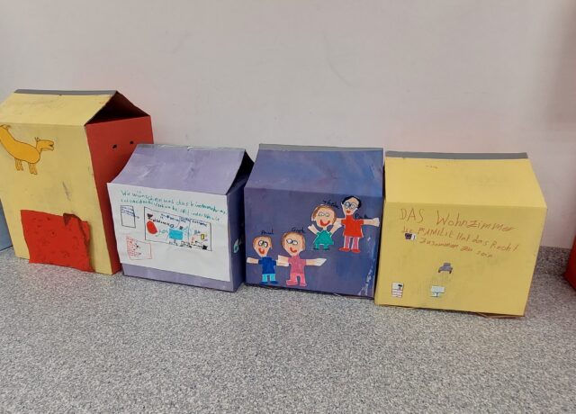 Bunt bemalte Kartons stehen nebeneinander. Auf den Kartons sind Kinderzeichnungen von Menschen und Kinderschrift, welche jedoch nicht lesbar ist.