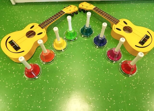 Zwei gelbe Ukulelen liegen auf grünem Boden, die Köpfe liegen eng aneinander. Acht Handglocken in sieben verschiedenen Farben stehen im Halbrund davor. Auf den Ukulelen sieht man einen Smily.