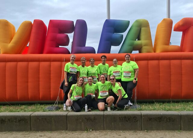 10 Läuferinnen und Läufer mit neongrünen T-Shirts stehen vor einem luftaufgeblasenen Schriftzug "Vielfalt". Das Wort strahlt in vielen verschiedenen Farben.