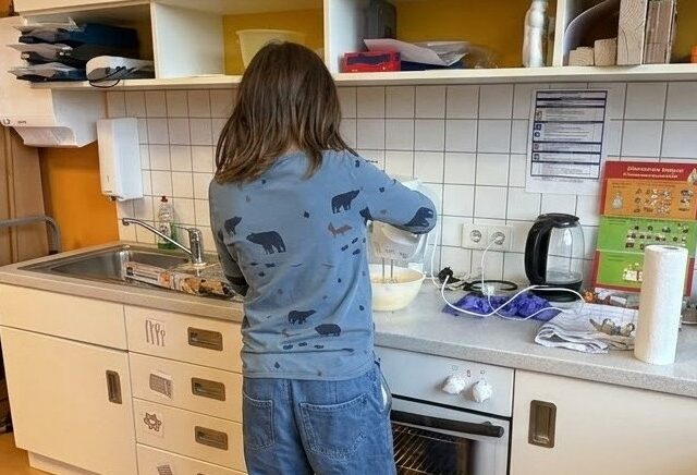 Ein Mädchen steht an einer Küchenzeile. Es ist von hinten zu sehen. Sie hat einen Handrührer in der Hand und bereitet einen Teig in einer weißen Schüssel zu. Im Umfeld sieht man weitere Küchenutensilien: Waschbecken, Wasserkoche, Küchenpapier, Handtücher und Backpapier.