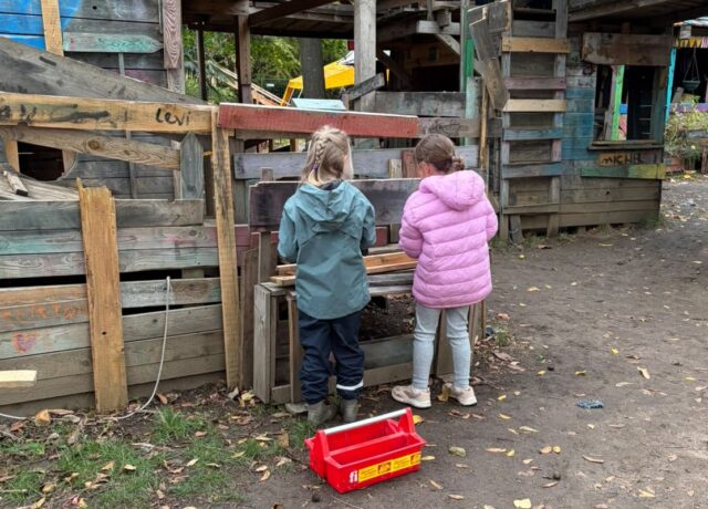 Zwei Kinder stehen in einem Abenteuerspielplatz vor einer Bretterwand. Sie scheinen etwas zu werken, man sieht sie jedoch von hinten und kann daher ihre Hände nicht sehen. Der Boden ist feucht, es scheint bewölkt zu sein.