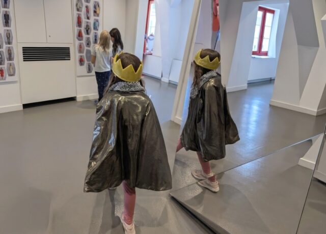 Ein Mädchen trägt eine gelbe Krone aus Stoff sowie einen graugrünen Umhang. Sie läuft an einem Spiegel vorbei, man sieht sie von hinten und teilweise im Spiegel. Im Hintergrund stehen zwei weitere Kinder, die Bilder an einer Wand anschauen.