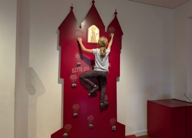 ein Mädchen in schwarzer Leggins und grauem T-Shirt klettert an einer roten Bolderwand. Das Kletterfeld ist wie ein Märchenturm gestaltet und steht vor einer weißen Zimmerwand.